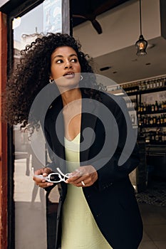 Beautiful young african girl looking away waving her hair stands near cafe during sunny spring day.