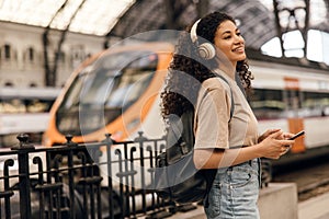 Beautiful young african girl stands on metro station enjoying her playlist on smartphone.