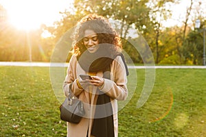 Beautiful young african happy woman walking outdoors in a spring park using mobile phone