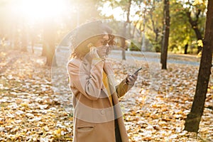 Beautiful young african happy woman walking outdoors in a spring park using mobile phone listening music with earphones