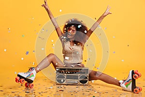 Beautiful young african woman with afro hairstyle throwing confetti, showing peace gesture while sitting in roller skates with bo