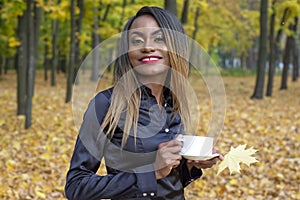 Beautiful young African woman drinking coffee from a white Cup on the background of autumn leaves in the Park