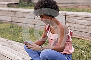 Beautiful young afro american woman using mobile phone, Sitting on wood stairs and smiling. wood background. Lifestyle outdoors