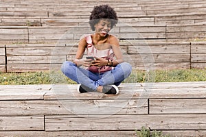 Beautiful young afro american woman using mobile phone, Sitting on wood stairs and smiling. wood background. Lifestyle outdoors