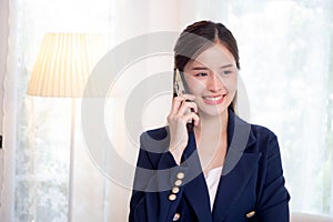 Beautiful young asian adviser or business woman talking on smartphone and smile while relax in the living room.