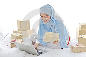 Beautiful and young asian arab woman in sleepwear with attractive look, sit on bed work with computer and online package box