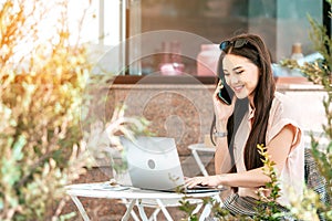 Beautiful young asian business woman talking cellphone while typing at the laptop.