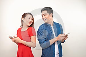 Beautiful young asian couple in casual clothes is using smartphones, looking at camera and smiling, standing back to back against