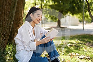 Beautiful young asian girl, student sits in park under tree and reading book, smiling, enjoying warm summer day outdoors