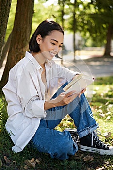 Beautiful young asian girl, student sits in park under tree and reading book, smiling, enjoying warm summer day outdoors