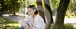 Beautiful young asian girl, student sits in park under tree and reading book, smiling, enjoying warm summer day outdoors