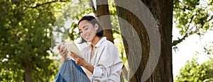 Beautiful young asian girl, student sits in park under tree and reading book, smiling, enjoying warm summer day outdoors