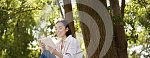 Beautiful young asian girl, student sits in park under tree and reading book, smiling, enjoying warm summer day outdoors