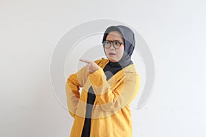 Beautiful young Asian Muslim woman in glasses, hijab and wearing yellow blazer with both hands pointing sideways and confused