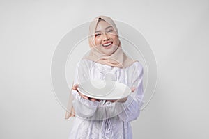 Beautiful young Asian Muslim woman in hijab presenting an empty plate with copy space over it, showing the food menu for iftar