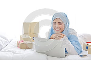 Beautiful and young asian muslim woman in sleepwear with attractive look, lies on bed with computer, mobile phone and online