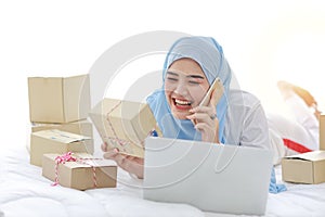 Beautiful and young asian muslim woman in sleepwear with attractive look, lies on bed with computer and online package box