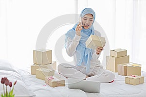 Beautiful and young asian muslim woman in sleepwear with attractive look, sit on bed with computer and online package box delivery photo