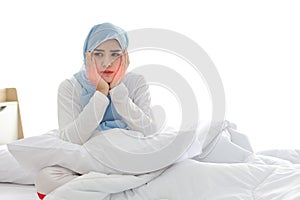 Beautiful and young asian muslim woman in sleepwear with tooth pain emotion sitting on bed. Cute girl touching cheek with her