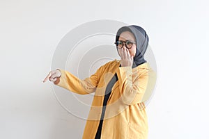 Beautiful young Asian Muslim woman, wearing glasses and yellow blazer covering mouth with hand and the other hand pointing