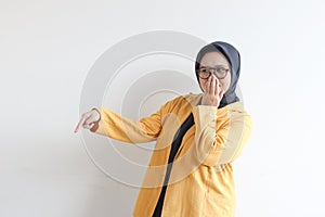 Beautiful young Asian Muslim woman, wearing glasses and yellow blazer covering mouth with hand and the other hand pointing