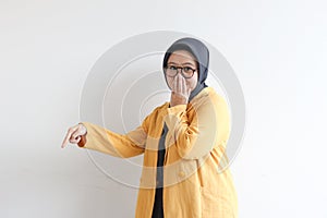 Beautiful young Asian Muslim woman, wearing glasses and yellow blazer covering mouth with hand and the other hand pointing