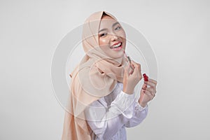 Beautiful young Asian Muslim woman wearing white dress and hijab putting on makeup applying lipstick. Fashion and cosmetics beauty