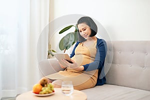 Beautiful young Asian pregnant woman using tablet and eating fruit when sitting on sofa