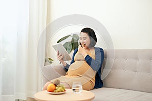 Beautiful young Asian pregnant woman using tablet and eating fruit when sitting on sofa