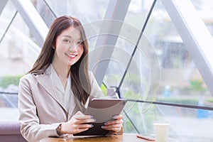 Beautiful young Asian professional business female in a cream-colored suit is looking at the camera while using tablet or notepad