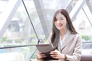 Beautiful young Asian professional business female in a cream-colored suit is looking at the camera while using tablet or notepad