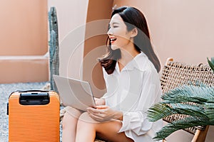 Beautiful young Asian solo traveling woman with smile using laptop. Female traveler sitting with orange suitcase and