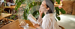 Beautiful young asian woman with a book in hands, sitting in cafe, drinking coffee and eating croissant, smiling