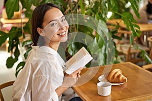 Beautiful young asian woman with a book in hands, sitting in cafe, drinking coffee and eating croissant, smiling