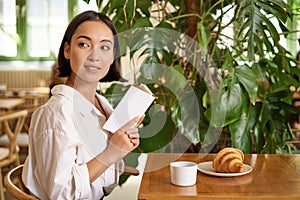 Beautiful young asian woman with a book in hands, sitting in cafe, drinking coffee and eating croissant, smiling