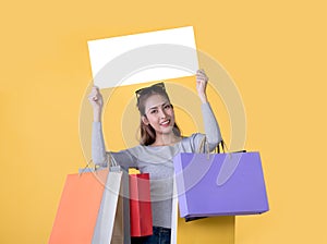 Beautiful young Asian woman carrying shopping bags and holding white banner with copy space isolated on yellow background