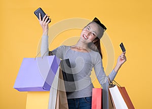 Beautiful young Asian woman with colourful shopping bags with smart phone and credit card isolated on yellow background