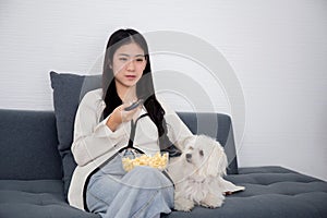 Beautiful young asian woman and dog sitting on sofa watching movie on television for leisure in living room.