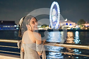 Beautiful young asian woman in dress smiling and sightseeing illuminated ferris wheel during cruise ship on the river in the night