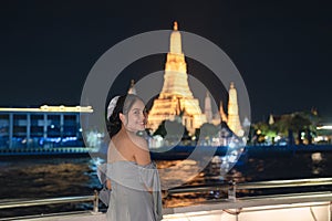 Beautiful young asian woman in dress standing and looking at camera on the deck with Wat Arun glowing in the night. Dinner cruise