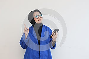 Beautiful young Asian woman in glasses, in hijab and wearing a blue blazer is holding a mobile phone and thinking