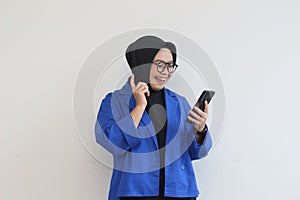 beautiful young asian woman in glasses, hijab and wearing a blue blazer looking at her mobile phone with happy face