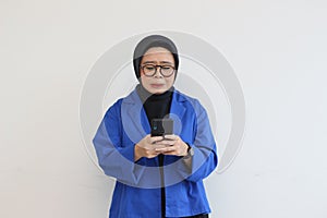 Beautiful young Asian woman in glasses, hijab and wearing a blue blazer looking at her mobile phone with a sad face