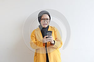 beautiful young asian woman in glasses, hijab and wearing yellow blazer looking at her mobile phone with shocked face