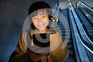 Beautiful young asian woman, going down an escalator, using smartphone, listening music in headphones while commuting to