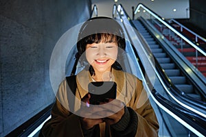 Beautiful young asian woman, going down an escalator, using smartphone, listening music in headphones while commuting to