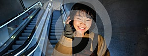 Beautiful young asian woman, going down an escalator, using smartphone, listening music in headphones while commuting to