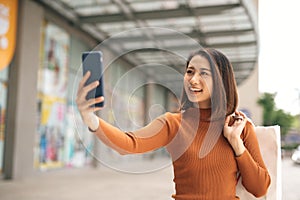 Beautiful young Asian woman holding phone to selfie with shopping bag outdoor shopping mall