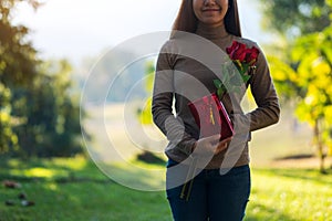 A beautiful young asian woman holding red roses flower and a gift box in the park