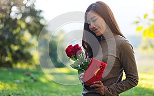 A beautiful young asian woman holding red roses flower and a gift box in the park